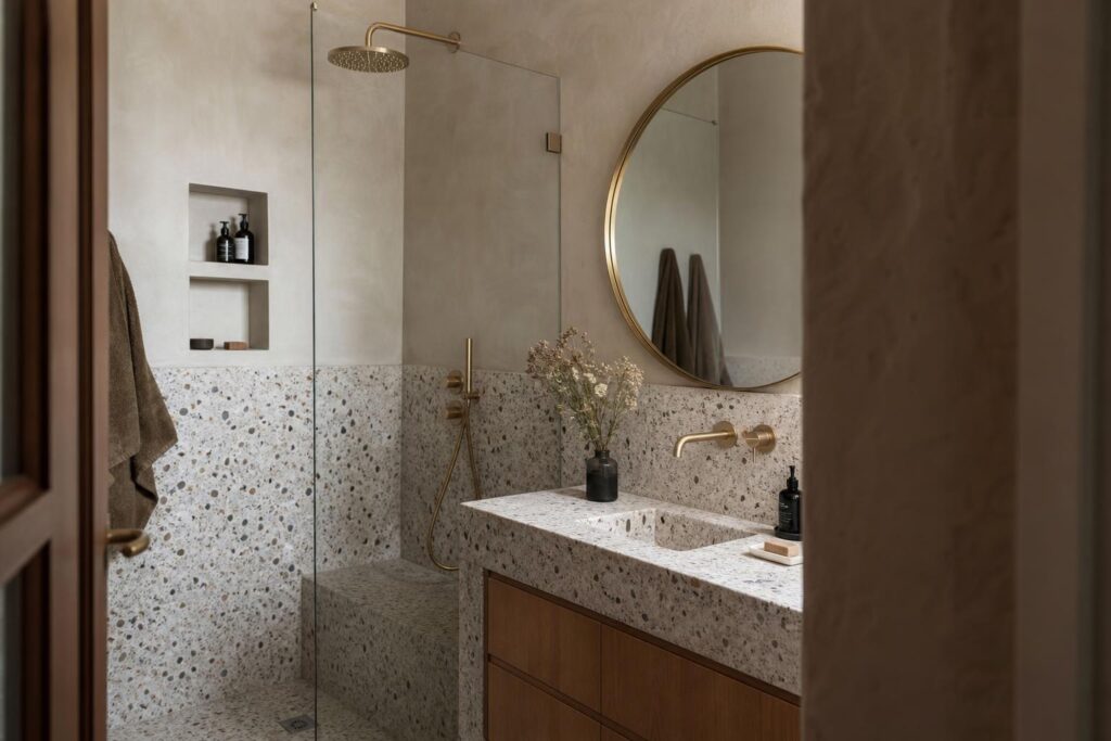 Petite salle de bain de 3 m² habillée de terrazzo et de tadelakt, avec robinetterie en laiton et miroir rond.